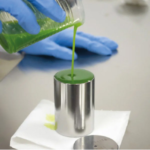 Pouring green ceramic ink from a glass container into a metal container, performed by an operator wearing a blue glove.